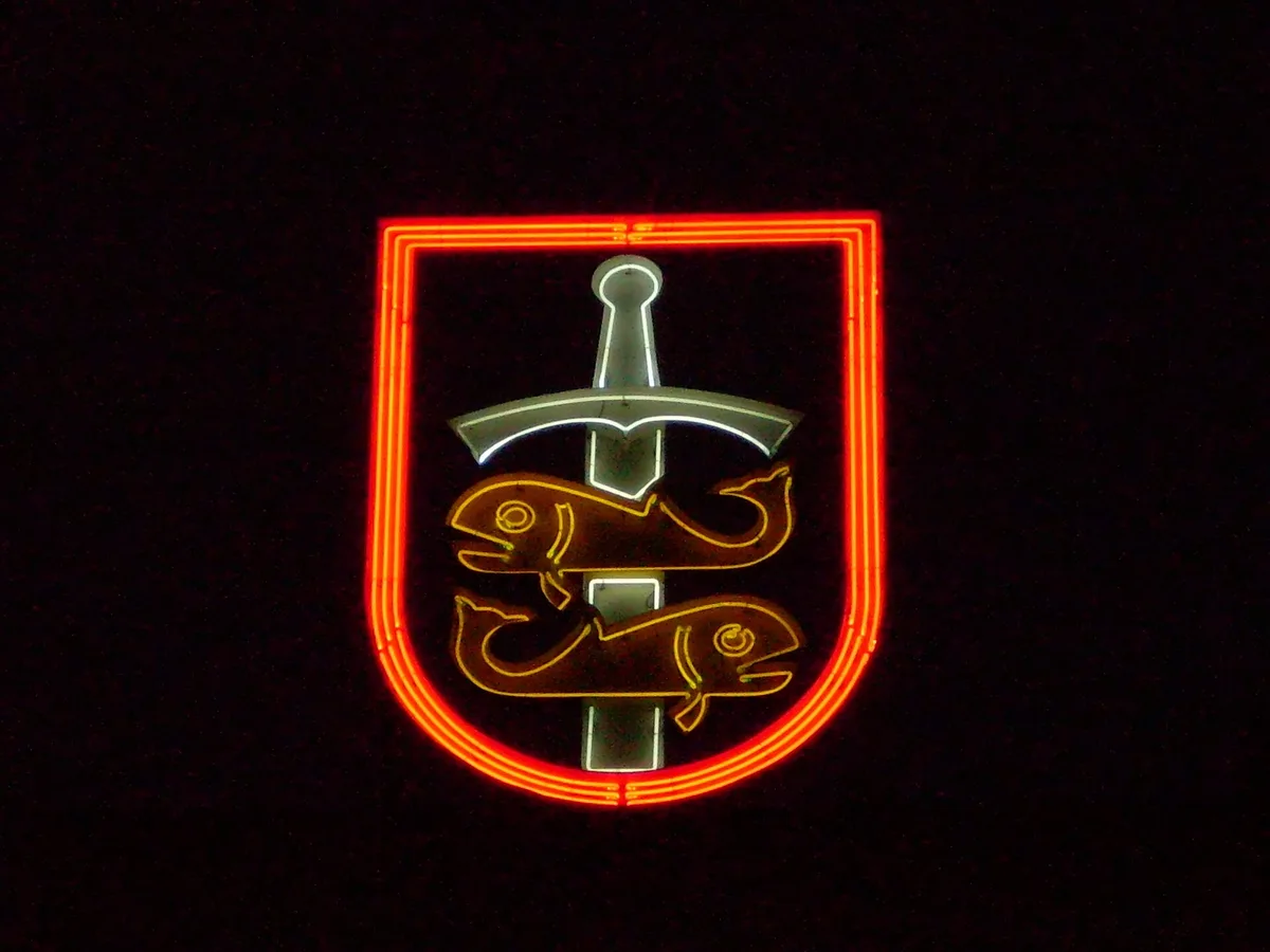 Neon w kształcie herbu Gdyni na ciemnym tle: czerwony obrys tarczy, w środku dwie żółte ryby przebite mieczem.