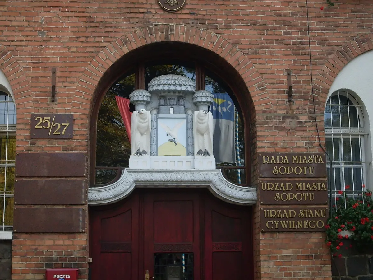 Ceglany portal wejściowy ratusza w Sopocie z tablicami urzędów; w zwieńczeniu, między dwiema figurami, tarcza z herbem miasta.